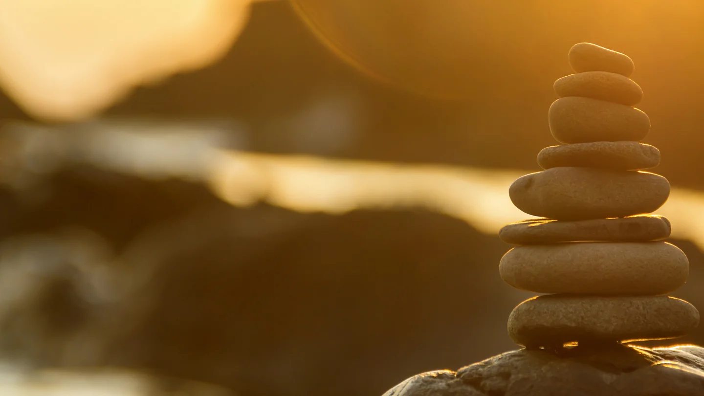 A stack of rough yet circular tan rocks is stacked in front of a rocky landscape, backed by blue sky, white streaking clouds, and yellow sunset.